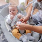 Handsome smiling young caucasian dad putting his adorable 6 months old son in baby rocker chair. Baby having confused facial expression. Looking away.